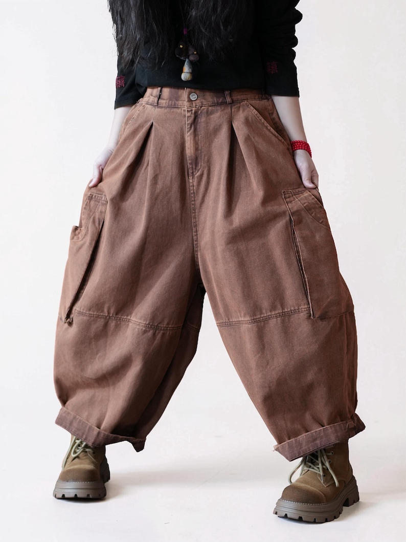May include: Brown cargo pants featuring a high-waisted design with front pleats, side cargo pockets, and a loose, wide-leg silhouette. The pants have a button closure, a zip fly, and cuffed hems, providing a relaxed fit with visible stitching.