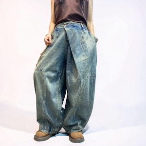 May include: Wide-leg denim trousers in a faded blue wash featuring an asymmetrical overlapping front panel design, a high-waisted fit with a button closure, and prominent vertical seam detailing running down the front of the loose-fitting legs.