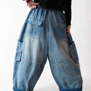 May include: Loose-fitting blue denim cargo pants featuring an elastic waistband, two large back pockets with flaps, and two side cargo pockets. The denim has a faded wash, a wide-leg silhouette, and thick, dark blue folded cuffs at the bottom hem.
