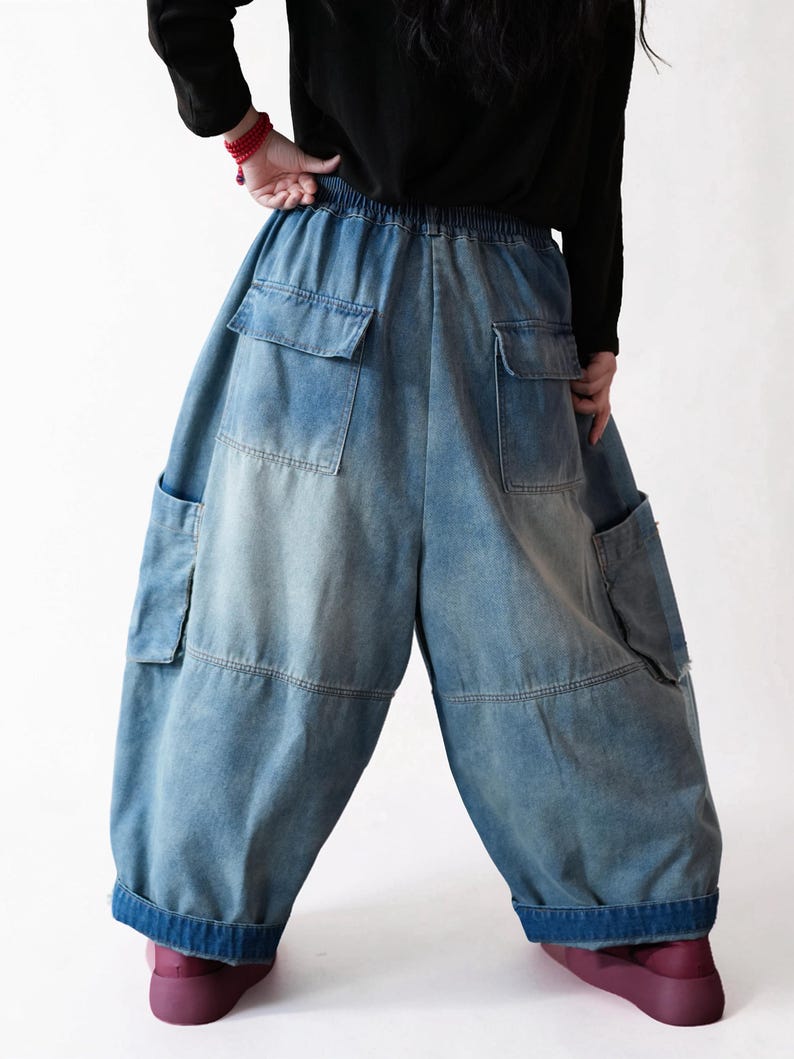May include: Loose-fitting blue denim cargo pants featuring an elastic waistband, two large back pockets with flaps, and two side cargo pockets. The denim has a faded wash, a wide-leg silhouette, and thick, dark blue folded cuffs at the bottom hem.