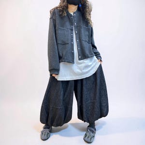 May include: A dark charcoal gray long-sleeve button-up jacket with two front pockets, worn over a long white undershirt. The outfit includes voluminous, wide-leg dark gray trousers with a gathered hem and gray athletic sneakers.