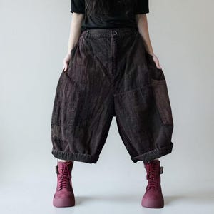 Op de afbeelding: Donkerbruine, wijd uitlopende cropped broek met een uniek patchwork design. De broek heeft grote zakken en wordt gecombineerd met bordeauxrode laarzen. De stof heeft een textuur en de broek heeft een knoopsluiting in de taille. De algehele stijl is casual en comfortabel.