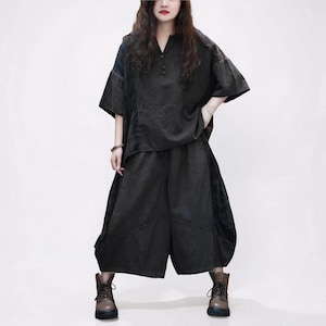 May include: A dark charcoal gray two-piece outfit consisting of a loose-fitting short-sleeved top with a button-up neckline and wide-leg cropped trousers. The ensemble features a textured fabric with subtle vertical paneling details on the sides.