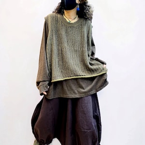 Könnte beinhalten: Eine ärmellose Strickweste in Olivgrün mit gerippter Textur, getragen über einem langärmeligen dunkelbraunen Hemd und einem dunkelbraunen Rock mit voluminöser, geraffter Silhouette. Das Ensemble besticht durch erdige Töne und abwechslungsreiche Stoffe.