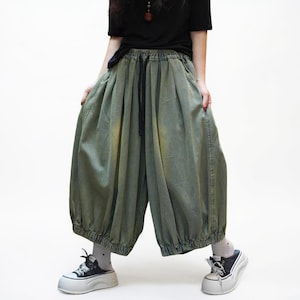 May include: Olive green wide-leg pants with a drawstring waist and elastic cuffs. The pants have a loose, baggy fit and are made of a soft, textured fabric. The model is wearing black and white sneakers.