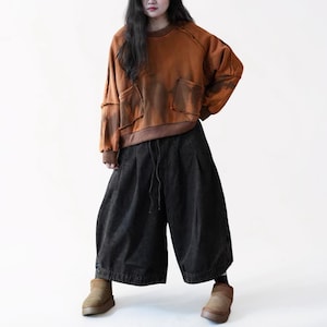 May include: A rust-colored sweatshirt with two front pockets and dark brown accents is paired with wide-leg, black culottes. The outfit is completed with brown ankle boots. The clothing has a relaxed, casual style.