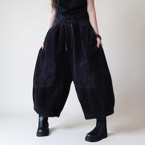 May include: Black corduroy pants with a wide, relaxed fit. The pants have an elastic waistband with a drawstring and side pockets. The model is wearing black boots. The pants are a dark, solid color.