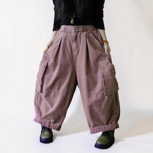 May include: Dusty rose wide-leg cargo pants featuring a high-waisted fit with front pleats, two large side cargo pockets, and a bright pink vertical stripe detail on the lower left leg. The pants have a button closure and a loose, baggy silhouette.