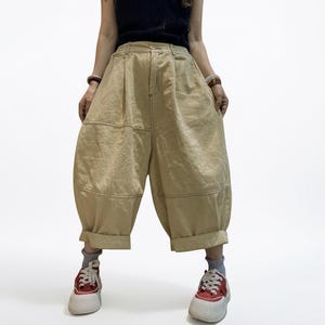 Può includere: Pantaloni beige a gamba larga con un design patchwork unico. I pantaloni hanno una vita alta e sono risvoltati alle caviglie. La modella indossa scarpe da ginnastica rosse e bianche. I pantaloni sono realizzati in un materiale robusto e presentano cuciture a vista.