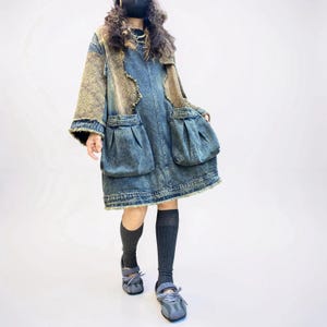 May include: A knee-length denim dress with a faded blue wash, featuring wide sleeves with brown patterned fabric panels, two large pleated front pockets, and a frayed hemline. The dress is paired with dark gray ribbed knee-high socks and gray shoes.