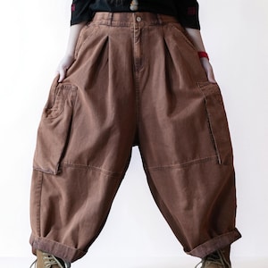 May include: Brown denim cargo pants featuring a high-waisted, pleated design with a loose, wide-leg silhouette. These pants include large side cargo pockets, a button and zipper fly closure, and cuffed hems, providing a relaxed and voluminous fit.