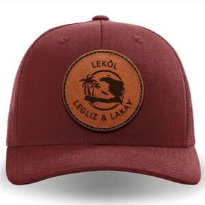 Op de afbeelding: Een bordeauxrode truckercap met een bruin leren patch met de omtrek van het eiland Hispaniola, palmbomen en de tekst "LEKÒL LEGLIZ & LAKAY".