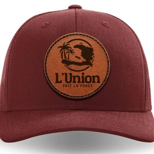 Pode incluir: Um boné de caminhoneiro de cor vinho com um remendo de couro marrom que apresenta uma palmeira e o contorno da ilha de Hispaniola. O remendo também inclui o texto "L'Union Fait La Force".
