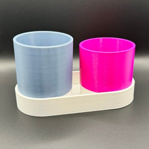 May include: Two cylindrical containers, one gray and one pink, are sitting on a white oval-shaped base. The containers are made of a smooth, textured material.