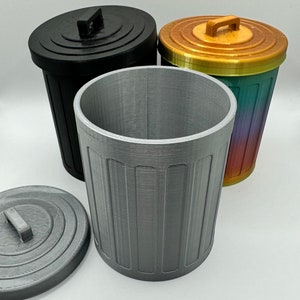 Pode incluir: Um conjunto de três latas de lixo decorativas em cores diferentes. Uma é prateada, outra preta e a terceira é um gradiente de ouro, verde e vermelho. Cada lata tem uma tampa e uma alça.
