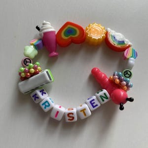 Puede incluir: Una pulsera colorida con varios dijes, incluyendo un batido rosa, un corazón arcoíris, un sol amarillo y una rebanada de sandía roja. La pulsera también presenta cuentas de letras blancas que deletrean "LISTEN".