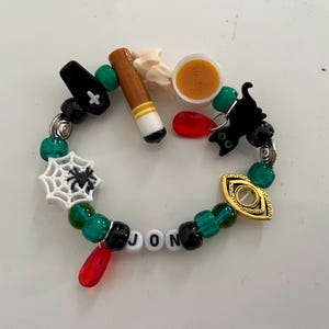May include: A Halloween-themed beaded bracelet. It includes green, black, and white beads, and charms such as a coffin, spiderweb, bat, and an eye. The name "JON" is spelled out with letter beads. The bracelet is a fun accessory.