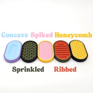 May include: Five oval soap dishes in different colours and textures. The designs include a light blue concave, a dark green spiked, a pink sprinkled, a red ribbed, and a yellow honeycomb. The words "Concave", "Spiked", "Honeycomb", "Sprinkled", and "Ribbed" are written above and below the dishes.