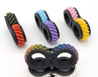 May include: A collection of colorful, 3D-printed gear-shaped fidget toys. Each toy features two interlocking gears in various color combinations, including green and red, blue and purple, and pink and yellow. The gears are set in a black frame.
