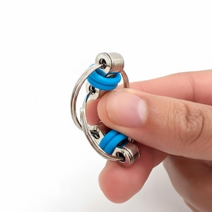 Op de afbeelding: Een zilveren metalen fidget-speeltje met twee verbonden secties. Elke sectie heeft een blauwe rubberen ring. Het speelgoed wordt in een hand gehouden tegen een witte achtergrond. Het speelgoed is ontworpen om met de vingers te worden gemanipuleerd.