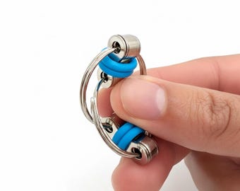 May include: A silver metal fidget toy with two connected sections. Each section has a blue rubber ring. The toy is held in a hand against a white background. The toy is designed to be manipulated with the fingers.