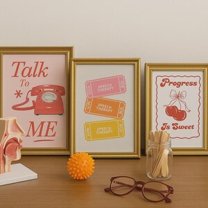 May include: Three framed prints with gold frames on a wooden surface. One print says "Talk To Me" with a red telephone graphic. Another has "Speech Therapy" tickets. The third says "Progress Is Sweet" with cherries.
