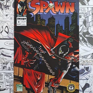 May include: Spawn comic book cover featuring a figure in a black suit and red cape, chained and holding chains. The title "Spawn" is in red and orange, with a skull graphic. The background shows a city skyline.