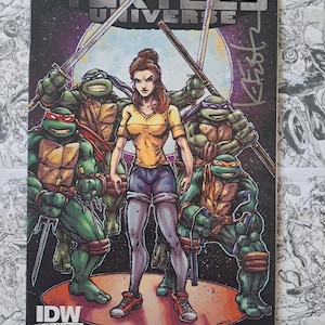 May include: Comic book cover for Teenage Mutant Ninja Turtles Universe #8. The cover features the four turtles with weapons, and a woman in a yellow shirt and denim shorts. The IDW Games Exclusive logo is in the bottom left corner.