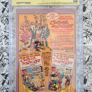 May include: A comic book cover featuring the characters from "Clever & Smart" in a German advertisement. The cover is encased in a protective holder. The advertisement includes text in German, promoting the video release of the comic.