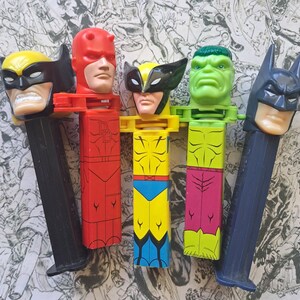 May include: A collection of five vintage PEZ dispensers featuring superhero characters. The dispensers include Wolverine, Daredevil, Hulk, and Batman. Each dispenser has a character head and a colorful body.