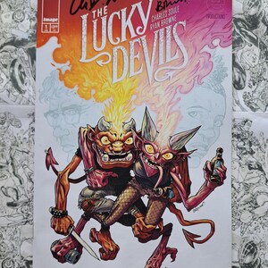 May include: Comic book cover for "The Lucky Devils" featuring two cartoon devils with horns, one holding a knife and the other a bottle. The title is in large, stylized letters with flames. Signed by the creators.