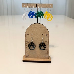 May include: A set of six paw print earrings on a wooden display stand. The earrings are made of acrylic and come in five colors: white, blue, green, yellow, and black.