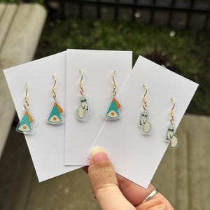 May include: Gold-colored dangle earrings with a whimsical design. Each earring features a clear, acrylic charm with a cartoonish figure in shades of blue, orange, and white. The earrings are displayed on white cards, showcasing the unique, artistic style.