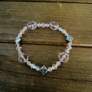 May include: A beaded bracelet featuring alternating light pink faceted crystal beads and clear smaller crystal beads, accented with silver-toned metal spacers and square-shaped floral patterned metal charms arranged in a circular pattern.
