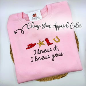 May include: A light pink crewneck sweatshirt featuring embroidered icons of a red cowboy hat, a yellow sheriff star, a red cowboy boot, and a gold horseshoe. Below the icons, the text reads, "I knew it, I knew you." A tag with a ladybug logo is attached.
