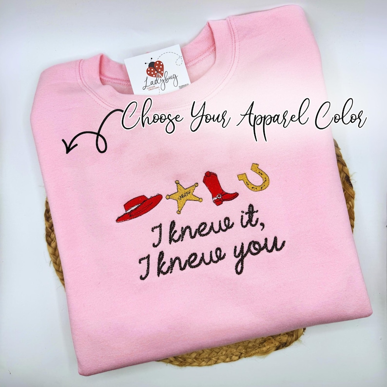 May include: A light pink crewneck sweatshirt featuring embroidered icons of a red cowboy hat, a yellow sheriff star, a red cowboy boot, and a gold horseshoe. Below the icons, the text reads, "I knew it, I knew you." A tag with a ladybug logo is attached.