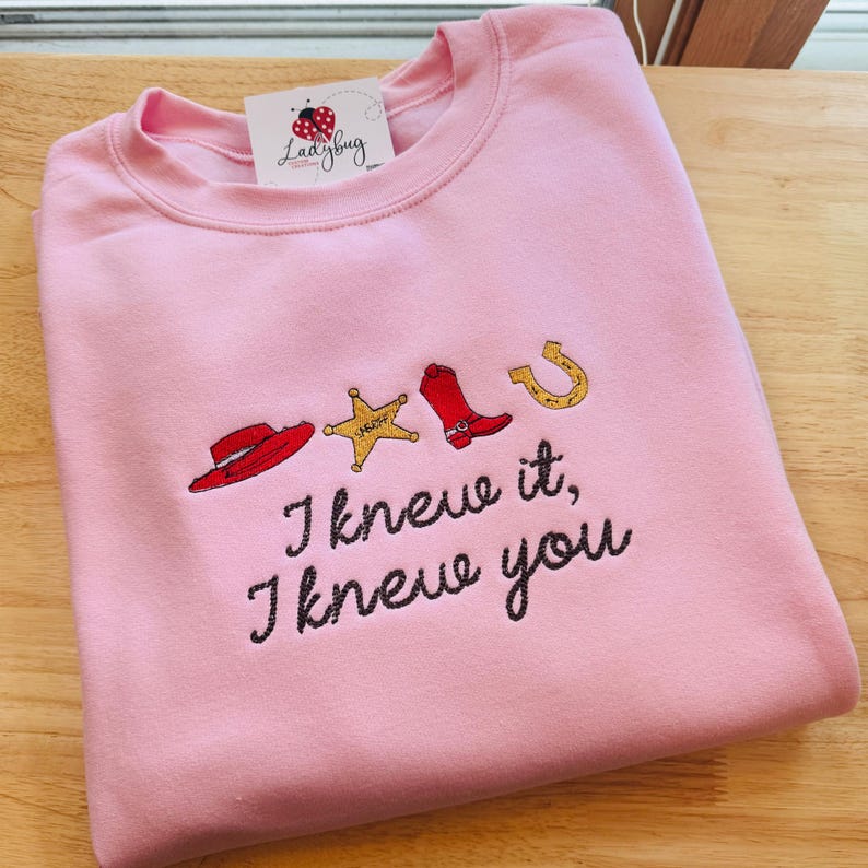 May include: A light pink crewneck sweatshirt featuring embroidered western-themed icons including a red hat, a yellow sheriff star, a red boot, and a gold horseshoe. Below the icons, the text reads, "I knew it, I knew you" in black.