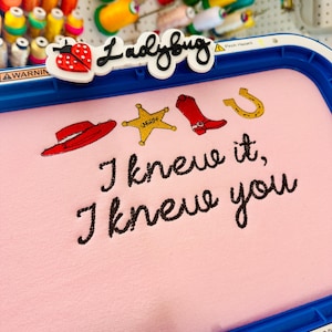 May include: A blue embroidery hoop holding light pink fabric with stitched designs of a red hat, a yellow sheriff star, a red cowboy boot, and a gold horseshoe. Below the icons, the black text reads, I knew it, I knew you.