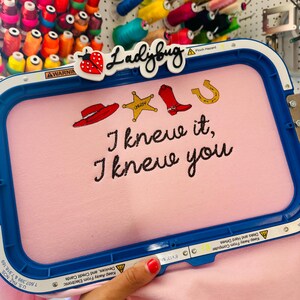 May include: A blue embroidery hoop holding light pink fabric with stitched designs of a red cowboy hat, a yellow sheriff star, a red cowboy boot, and a gold horseshoe. The text "I knew it, I knew you" is embroidered in black thread below the icons.