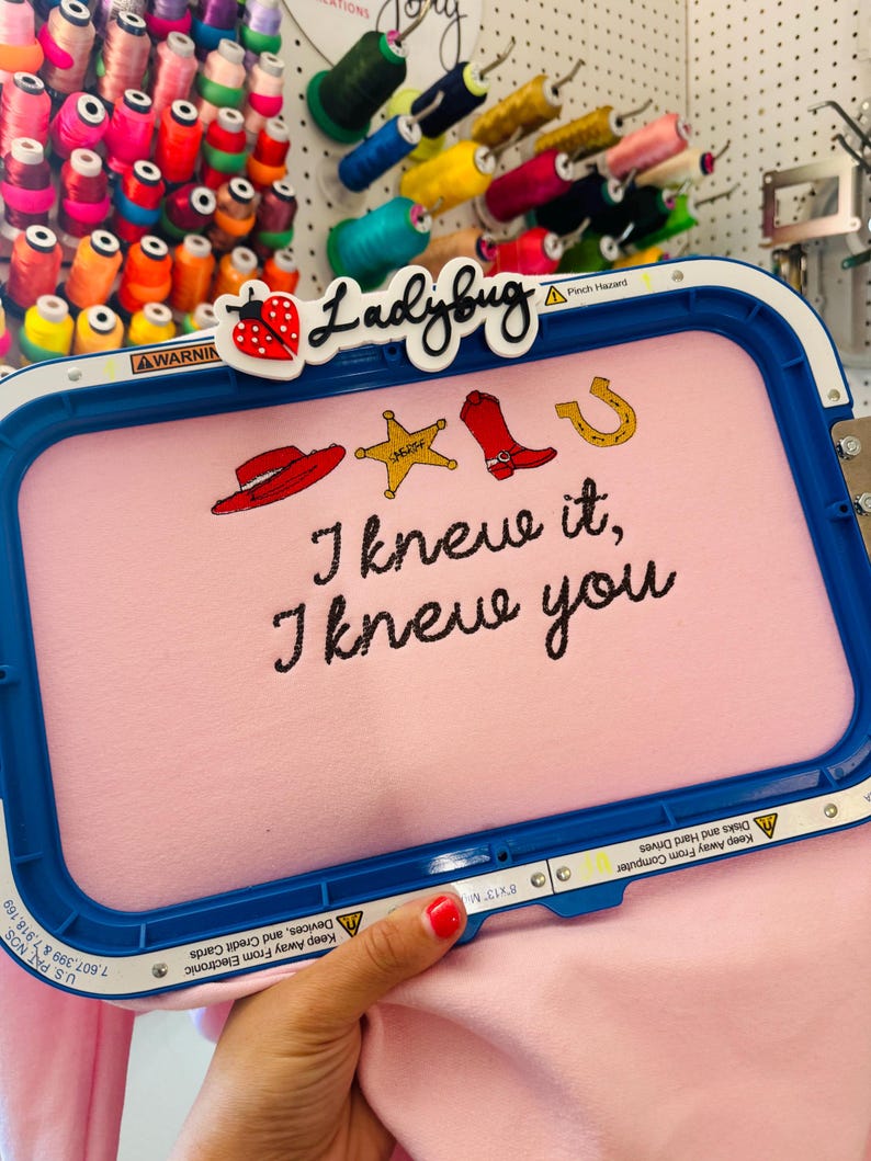 May include: A blue embroidery hoop holding light pink fabric with stitched designs of a red cowboy hat, a yellow sheriff star, a red cowboy boot, and a gold horseshoe. The text "I knew it, I knew you" is embroidered in black thread below the icons.