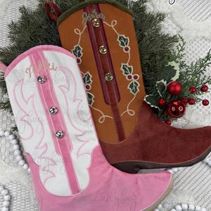 May include: Two decorative cowboy boot Christmas stockings. One is pink and white with silver accents, the other is brown with green and red accents. The boots are personalized with names and holly embroidery. The text "Embroidered Personalization" is at the top.