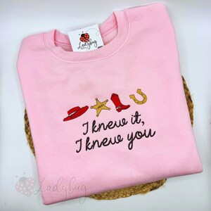 May include: A light pink crewneck sweatshirt featuring an embroidered design of a red cowboy hat, a gold sheriff star, a red cowboy boot, and a gold horseshoe. Below the icons, the text reads, "I knew it, I knew you" in black cursive.