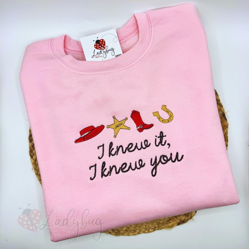 May include: A light pink crewneck sweatshirt featuring an embroidered design of a red cowboy hat, a gold sheriff star, a red cowboy boot, and a gold horseshoe. Below the icons, the text reads, "I knew it, I knew you" in black cursive.