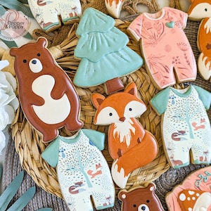 May include: An assortment of decorated sugar cookies in a woodland theme. The cookies include onesies with forest scenes, foxes, bears, and a Christmas tree. The cookies are decorated with brown, orange, green, and white icing.