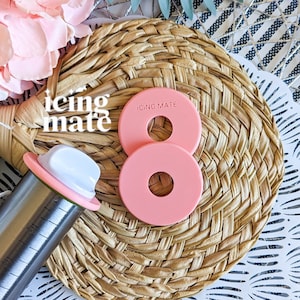 May include: Two pink plastic icing mate rings with the text "ICING MATE" on the top ring. The rings are on a woven straw mat. A silver piping tool with a pink and white top is in the background.