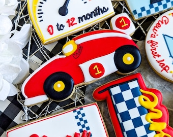 May include: A set of decorated cookies for a first birthday party with a racing car theme. The cookies feature a red race car, a checkered flag, a speedometer with the text "0 to 12 months", and a cookie with the word "Crew".