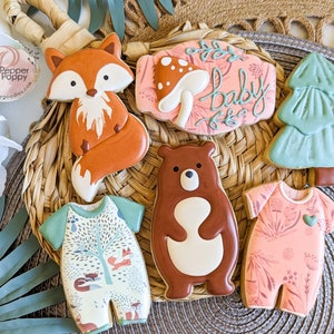 Pu&ograve; includere: Una collezione di biscotti di zucchero decorati a tema bosco. I biscotti includono una volpe, un orso, tutine per bambini, un fungo, un albero e un cartello con la scritta "baby". I biscotti sono decorati con glassa arancione, marrone, verde e rosa.