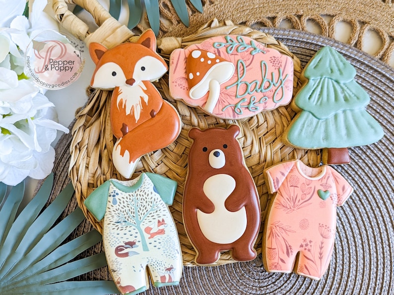 Pu&ograve; includere: Una collezione di biscotti di zucchero decorati a tema bosco. I biscotti includono una volpe, un orso, tutine per bambini, un fungo, un albero e un cartello con la scritta "baby". I biscotti sono decorati con glassa arancione, marrone, verde e rosa.