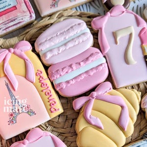 May include: Assortment of decorated sugar cookies in various shapes, including macarons, perfume bottles, and a dress. The cookies are primarily pink, yellow, and white, with decorative bows and Eiffel Tower imagery. The cookies are arranged on a woven surface.