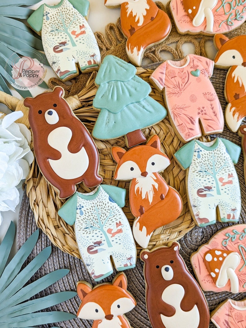 Pu&ograve; includere: Un assortimento di biscotti decorati a tema bosco. I biscotti includono tutine con scene di foresta, volpi, orsi e un albero di Natale. I biscotti sono decorati con glassa marrone, arancione, verde e bianca.